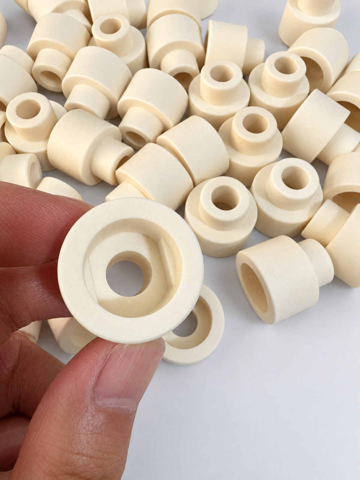 A pile of cream stepped ceramic bushings with through bores; a hand holds one to show the counterbore in its larger end.