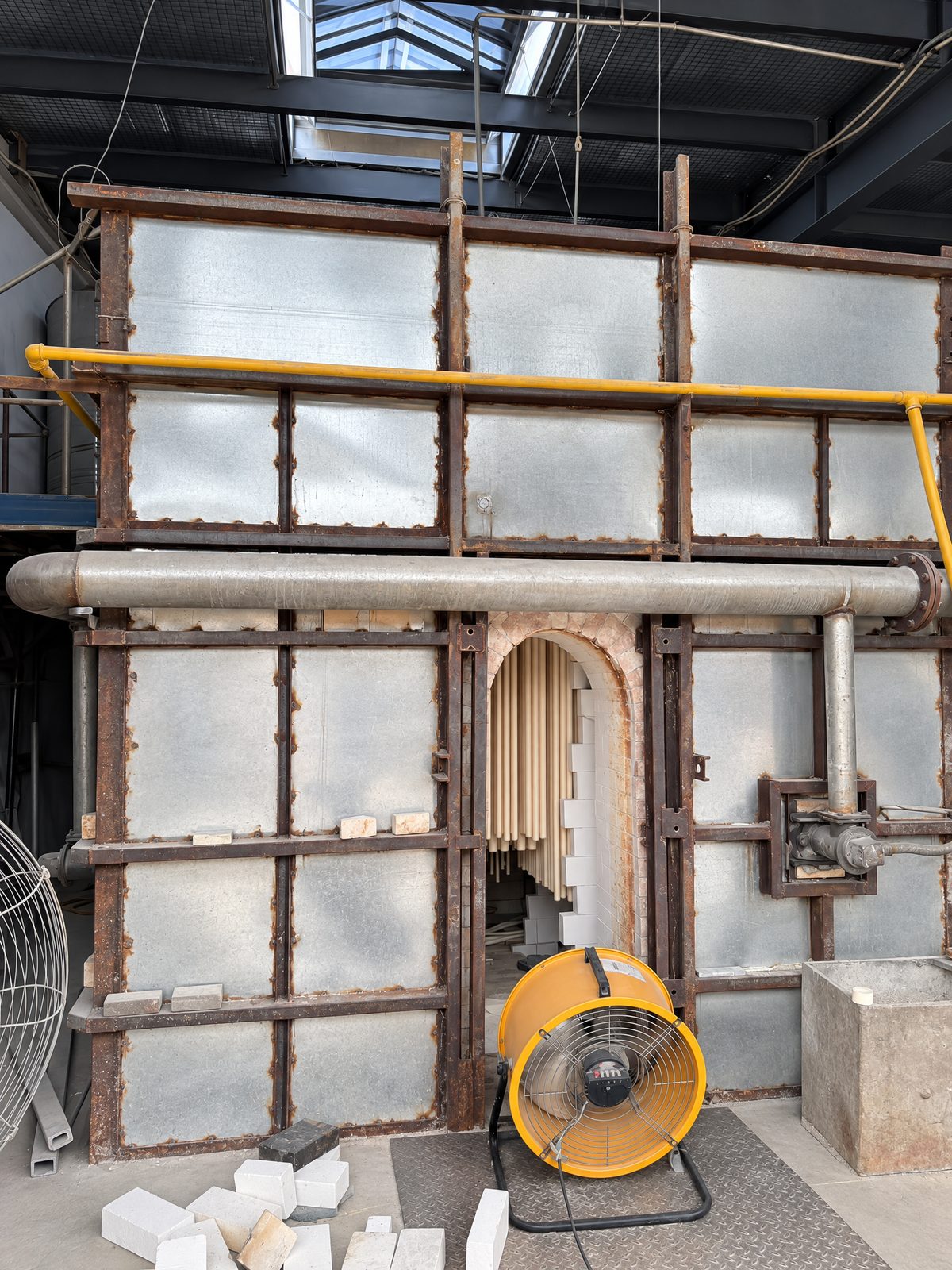 A large gas-fired kiln with a steel-framed front; through the arched door, long ceramic tubes hang inside.
