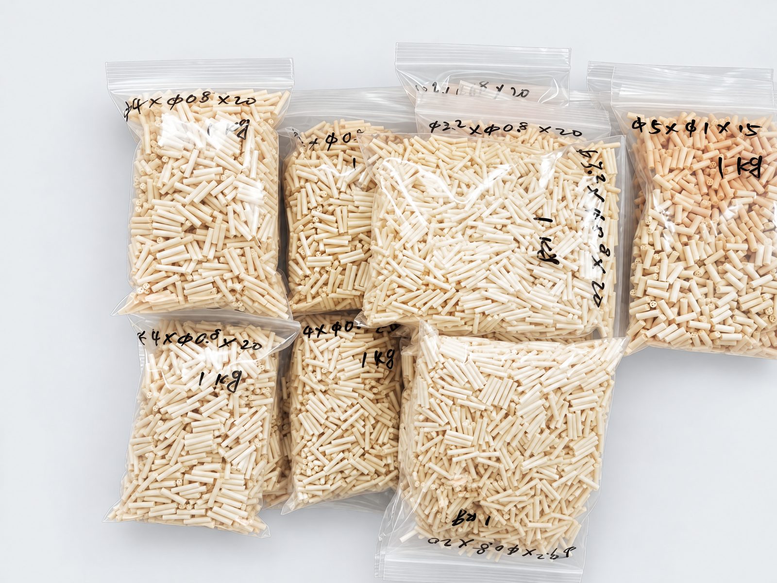 Zip-lock bags of short cream multi-bore beads, each marked by hand with its size and 1 kg, on a plain grey background.