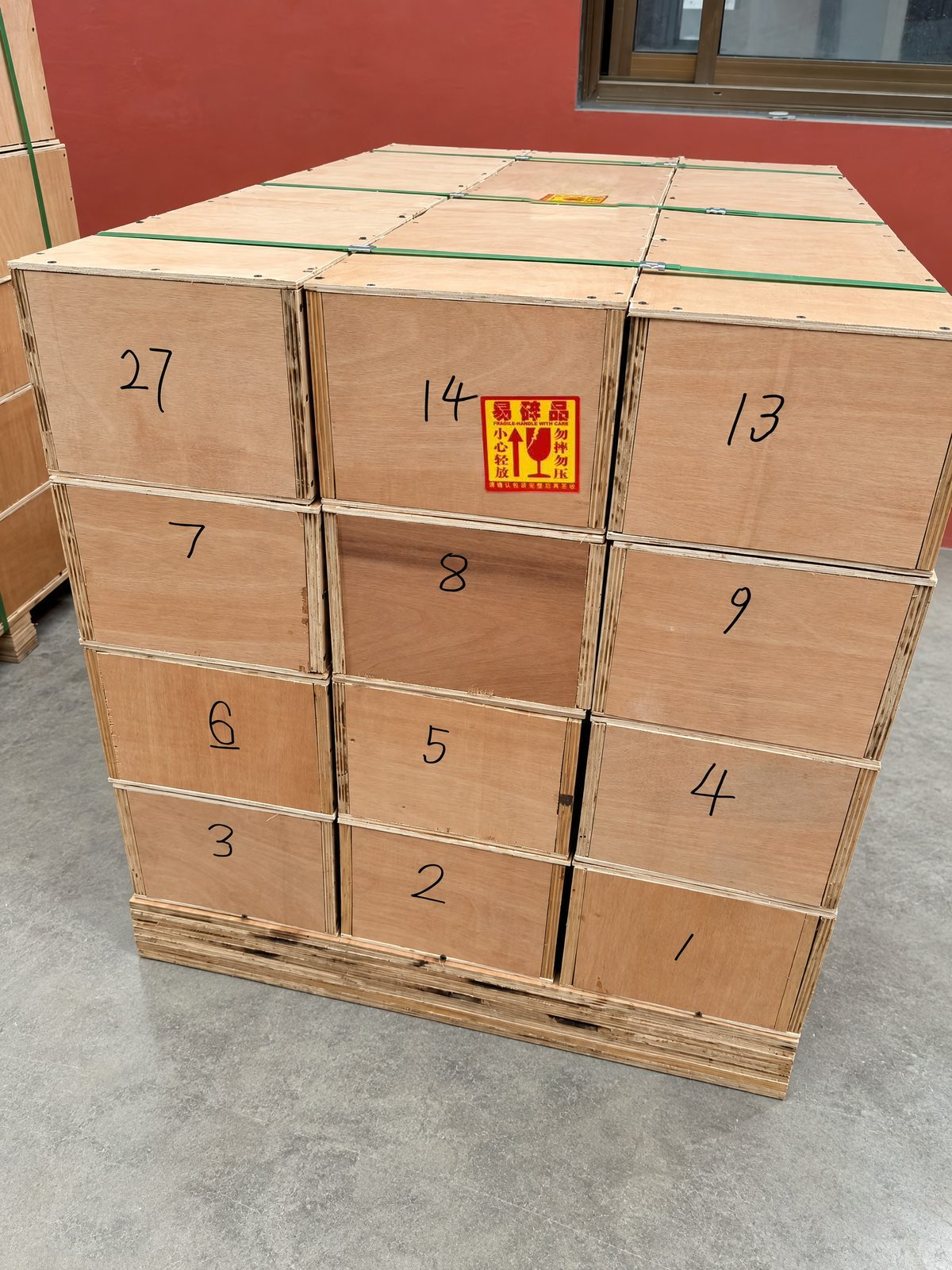 Twelve numbered plywood crates stacked three wide and four high on a pallet, strapped with green bands, with a fragile sticker on one crate.